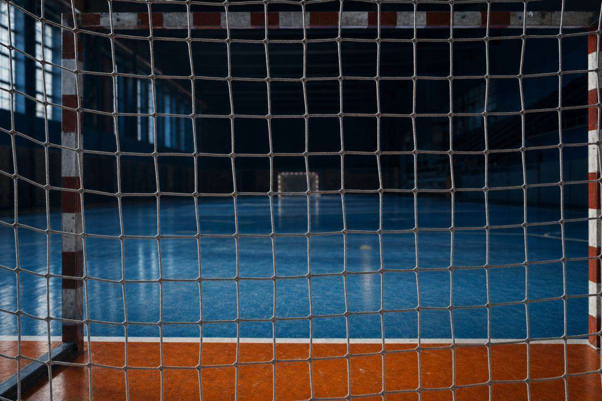 Blick durch das Netz eines Handballtors auf ein leeres blaues Hallenfeld