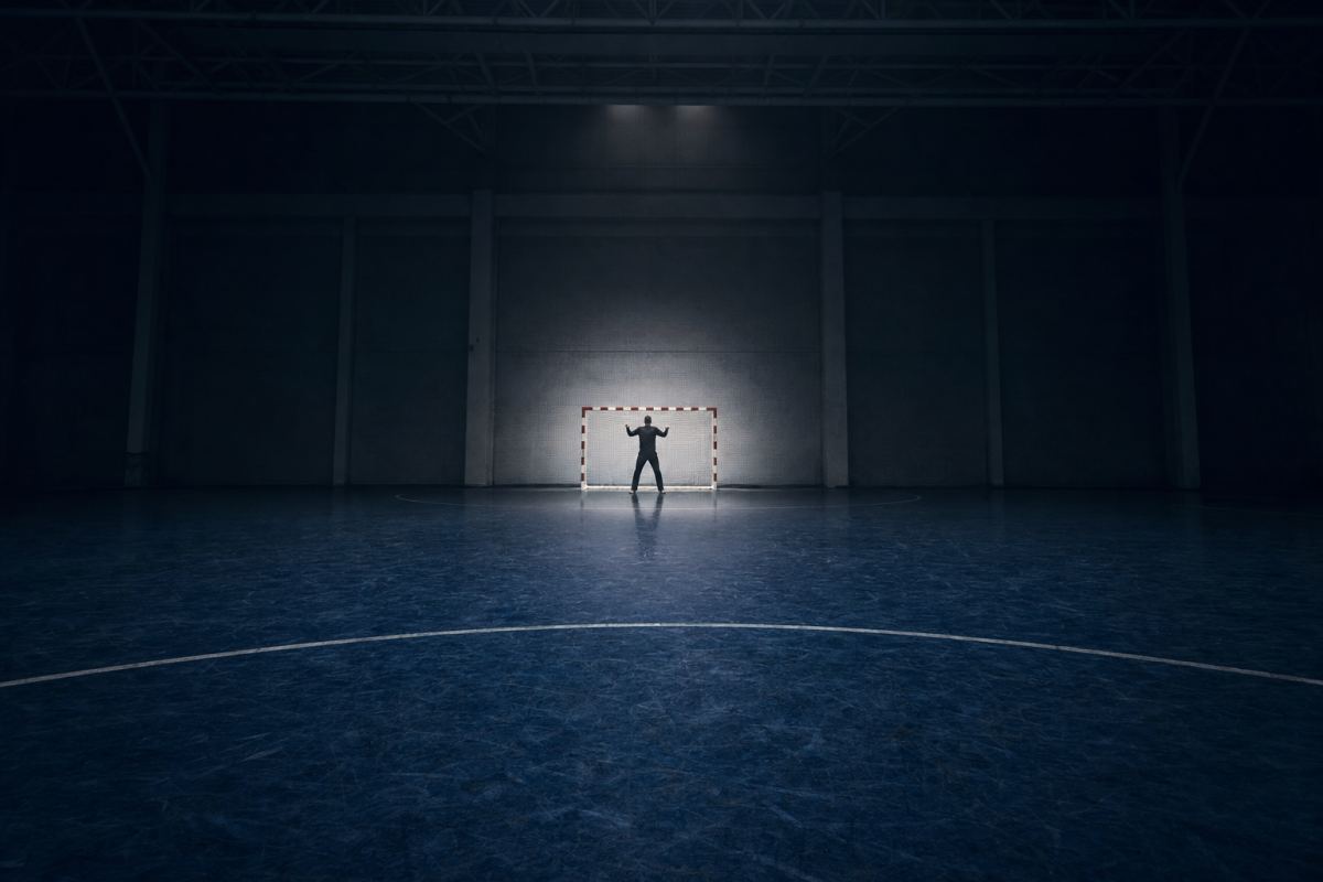Torwart allein im Tor einer leeren Handballhalle, von der Mittellinie aus gesehen