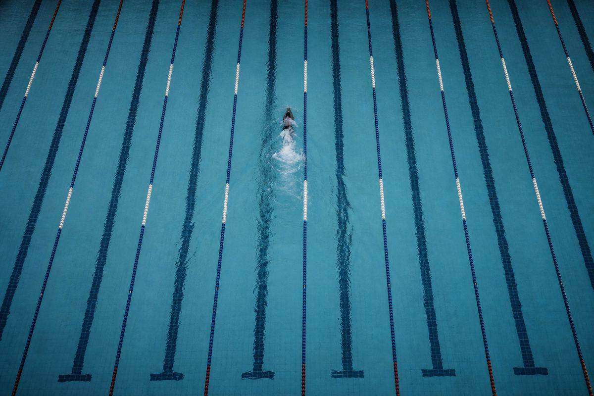 Wettkampfbecken von direkt oben fotografiert, Bahnbegrenzungen als strenges Muster, in einer Bahn ein Schwimmer als Wasserwirbel