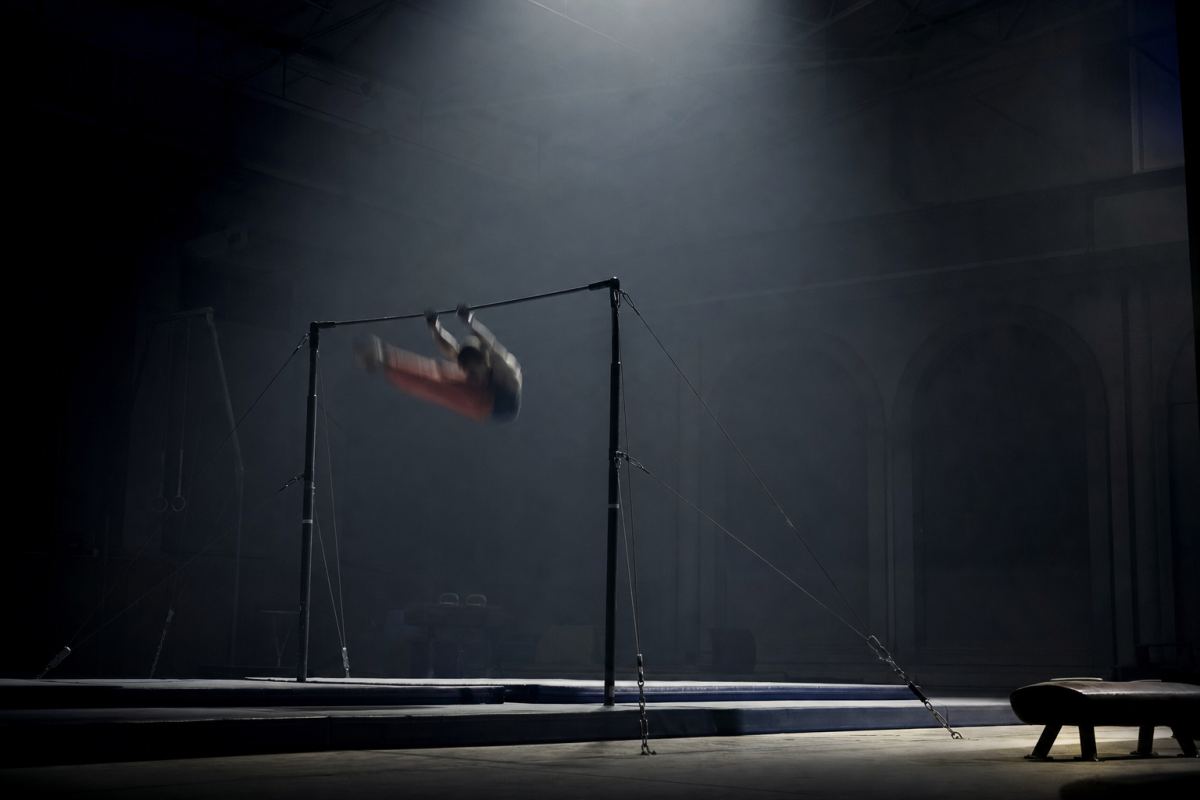 Weite dunkle Turnhalle, in der Mitte ein Reck, darüber ein Turner in der Flugphase als kleine Silhouette