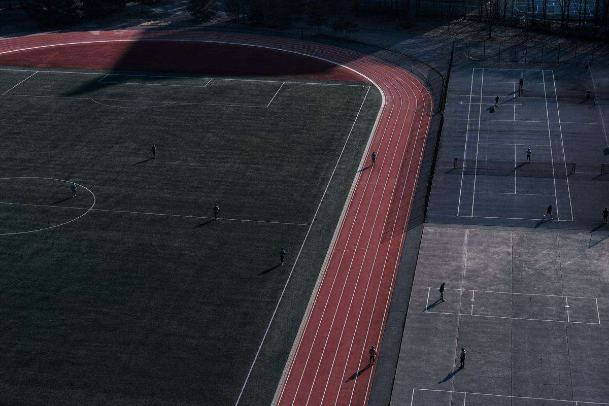 Kommunale Sportanlage aus großer Höhe: Laufbahn, Rasenplatz und zwei Hartplätze nebeneinander, darauf verteilt kleine entfernte Figuren