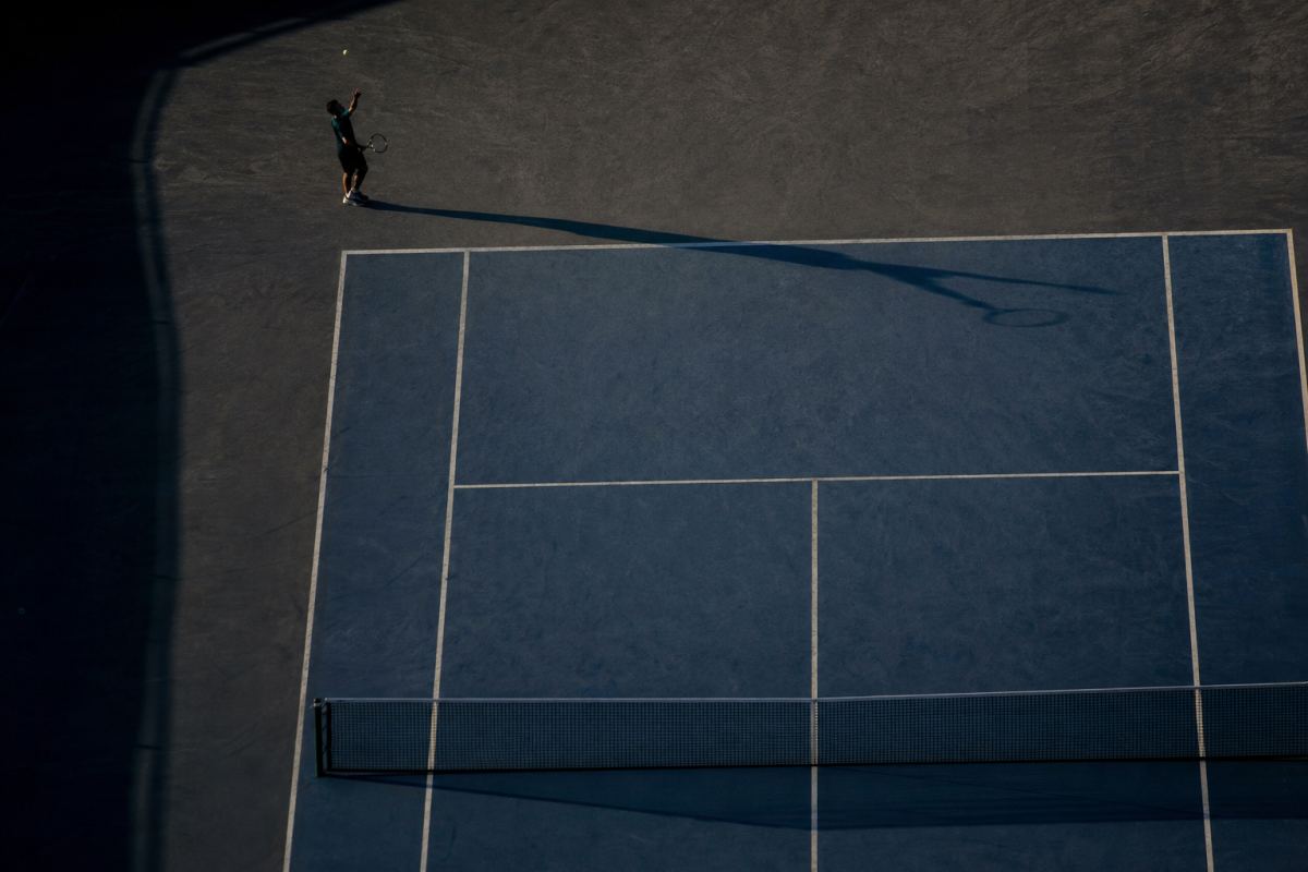 Tennisplatz von hoch oben in tiefer Sonne, der lange Schatten eines einzelnen Spielers reicht quer über den Platz